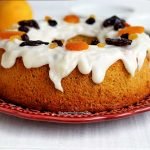 Bolo redondo com furo no meio decorado com cobertura branca que cai pela lateral com frutas secas no topo sobre prato cor de vinho em uma mesa branca