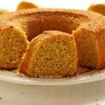 Bolo de laranja redondo com furo no meio partido sobre um prato branco, no meio do bolo tem uma fatia mostrando a textura aerada da massa, ao fundo laranjas desfocadas em uma mesa branca