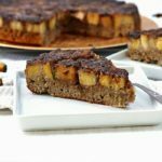 Fatia triangula de bolo de banana caramelizado na superfície, ao lado mais uma fatia de bolo e ao fundo o bolo partido em uma mesa branca