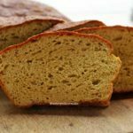 Pão caseiro fatiado, são três fatias apoiadas no pão sobre uma tábua de madeira em uma mesa branca