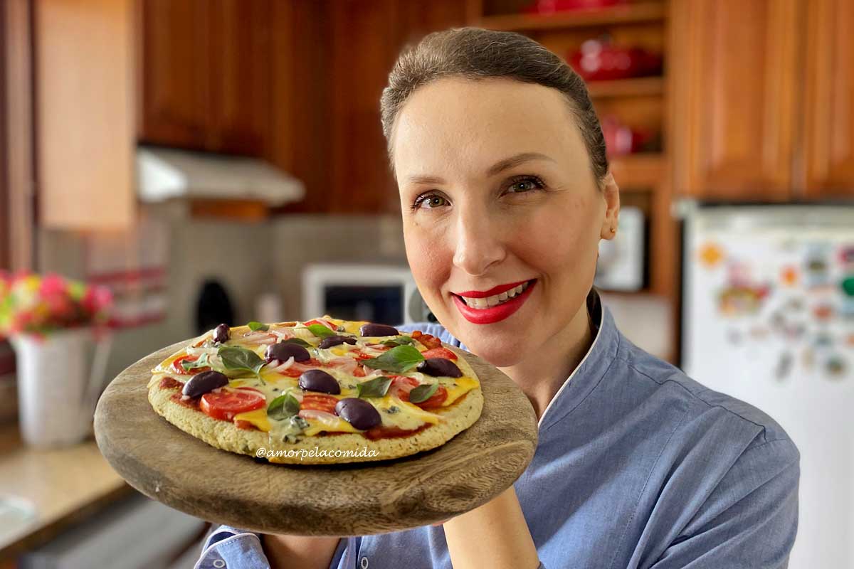 Mulher loira em uma cozinha com uniforme azul de chef segurando uma tábua de madeira, sobre a tábua a pizza de frigideira