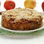 Torta de maçã redonda com cobertura de flocos finos de aveia sobre mesa branca com maçãs ao fundo