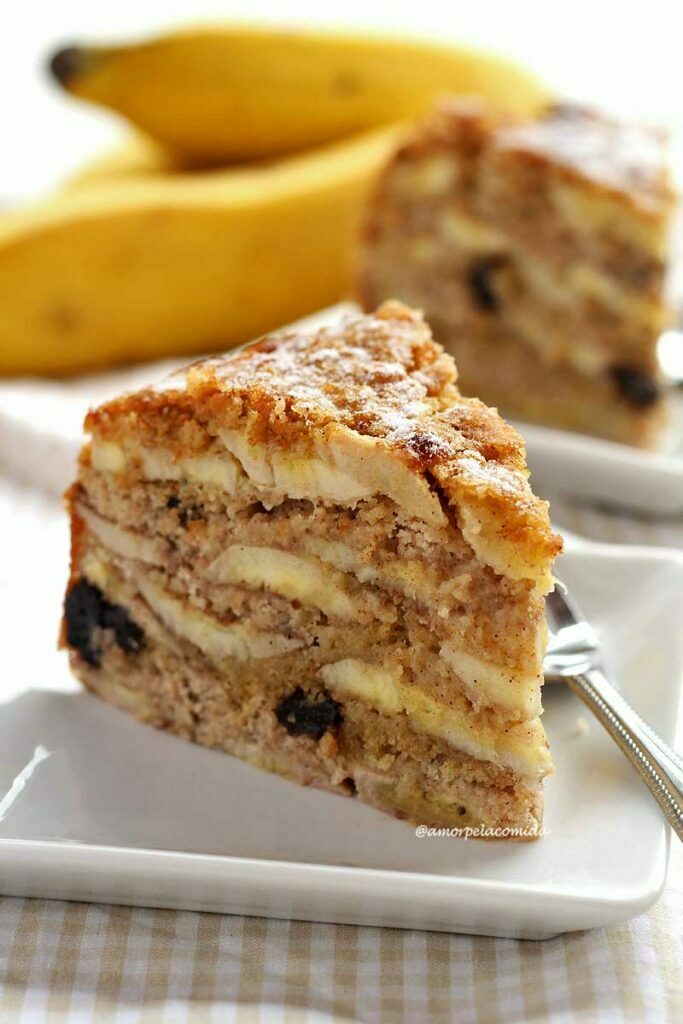 Fatia de torta de banana sobre prato quadrado branco pequeno, ao fundo mais um pedaço triangular e bananas desfocadas