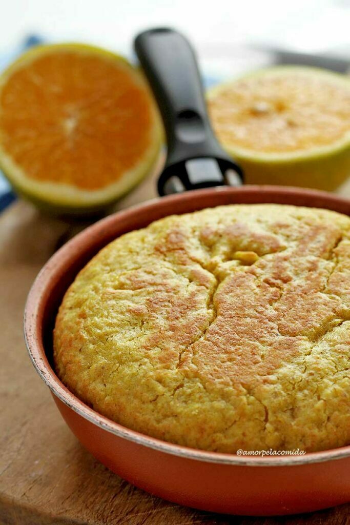 Bolo de laranja dentro de uma frigideira pequena sobre tábua de madeira de cor clara ao fundo 1 laranja partida ao meio
